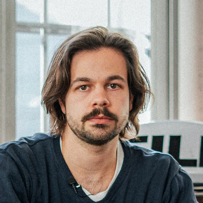 Tom Beckers — Le Werk, Köln Tom Beckers, Producer und Content Creator bei Le Werk in Köln, mit schulterlangem Haar und Schnurrbart vor einem hellen Fenster