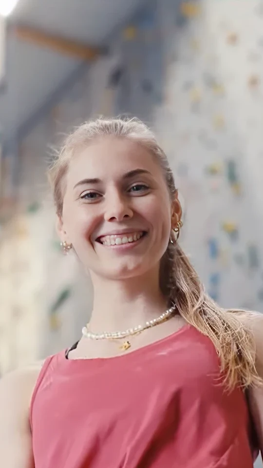 Standbild aus dem Athletenporträt für Allianz × Olympia: Kletterin Hannah Meul lacht in die Kamera, hinter ihr unscharf die bunte Boulderwand